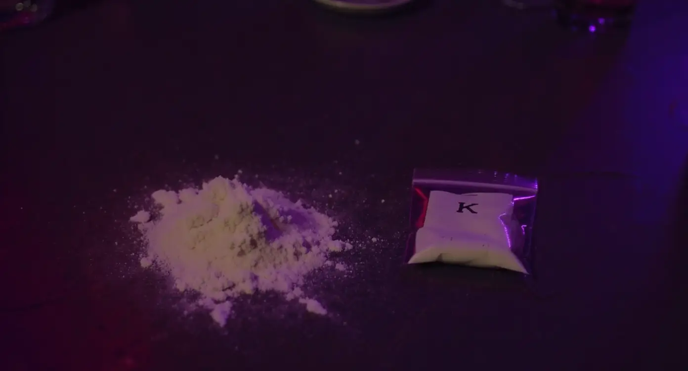 Hvidt ketaminpulver og plastikpose mærket K ved siden af et drinksglas på et mørkt festbord